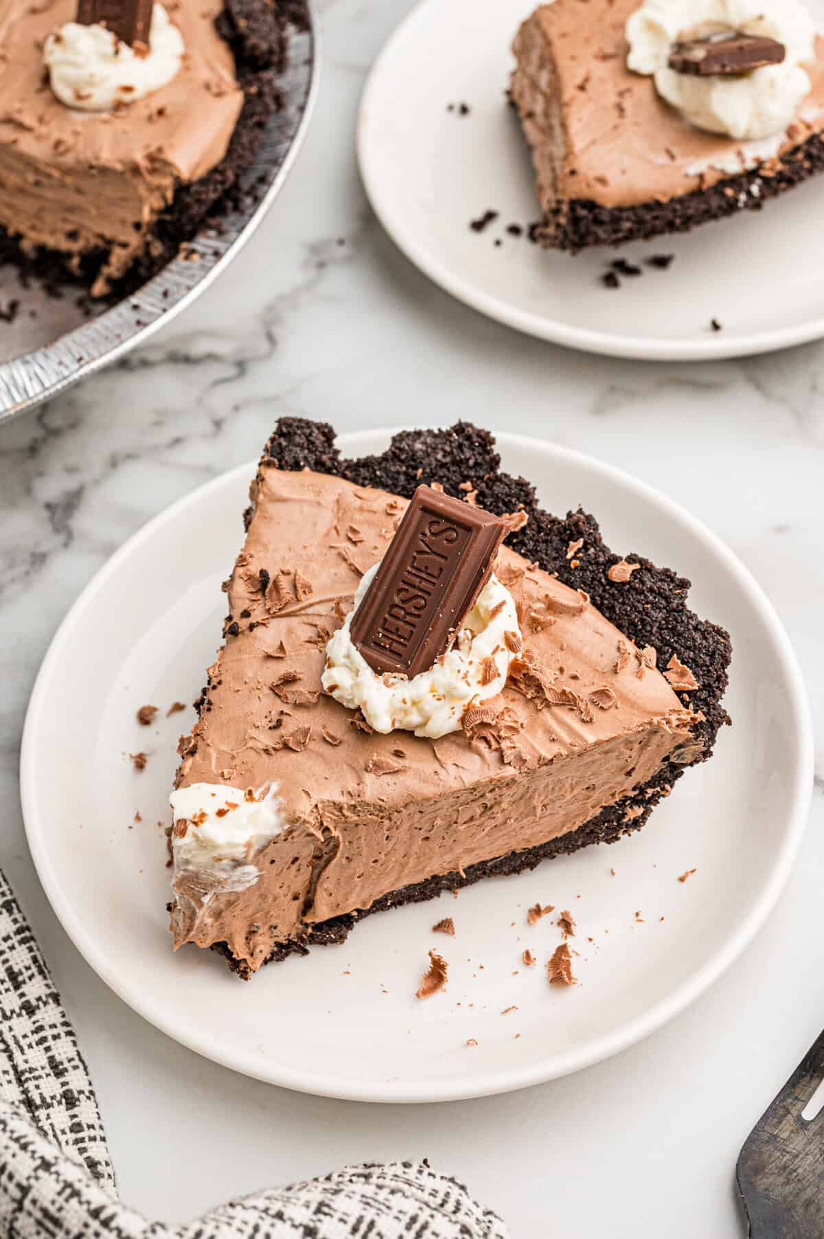 A slice of chocolate pie on a white plate with a second plated slice and the pie behind it.
