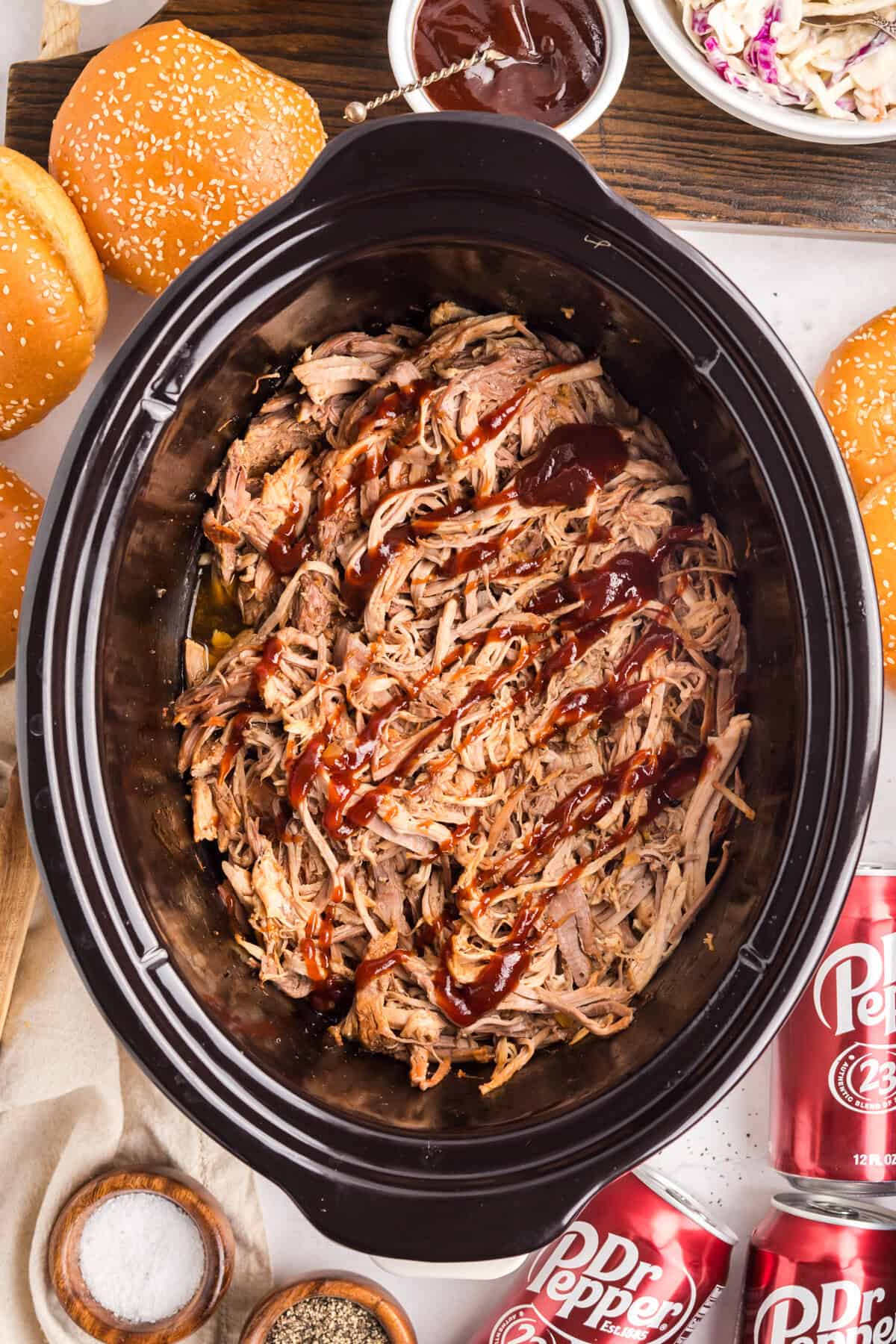 Overhead of a slow cooker full of shredded pork drizzled with barbecue sauce, sesame buns and Dr Pepper cans nearby.