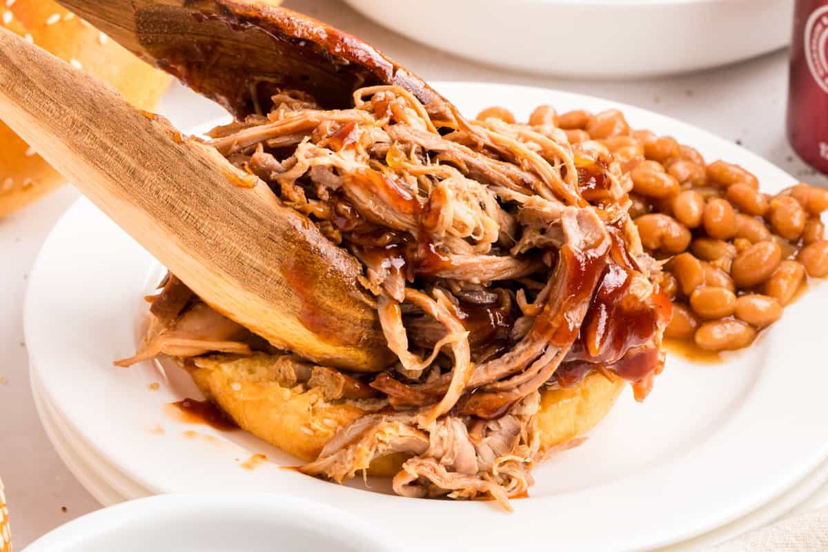 Wooden tongs piling barbecue pulled pork onto a sesame bun with baked beans on a white plate.