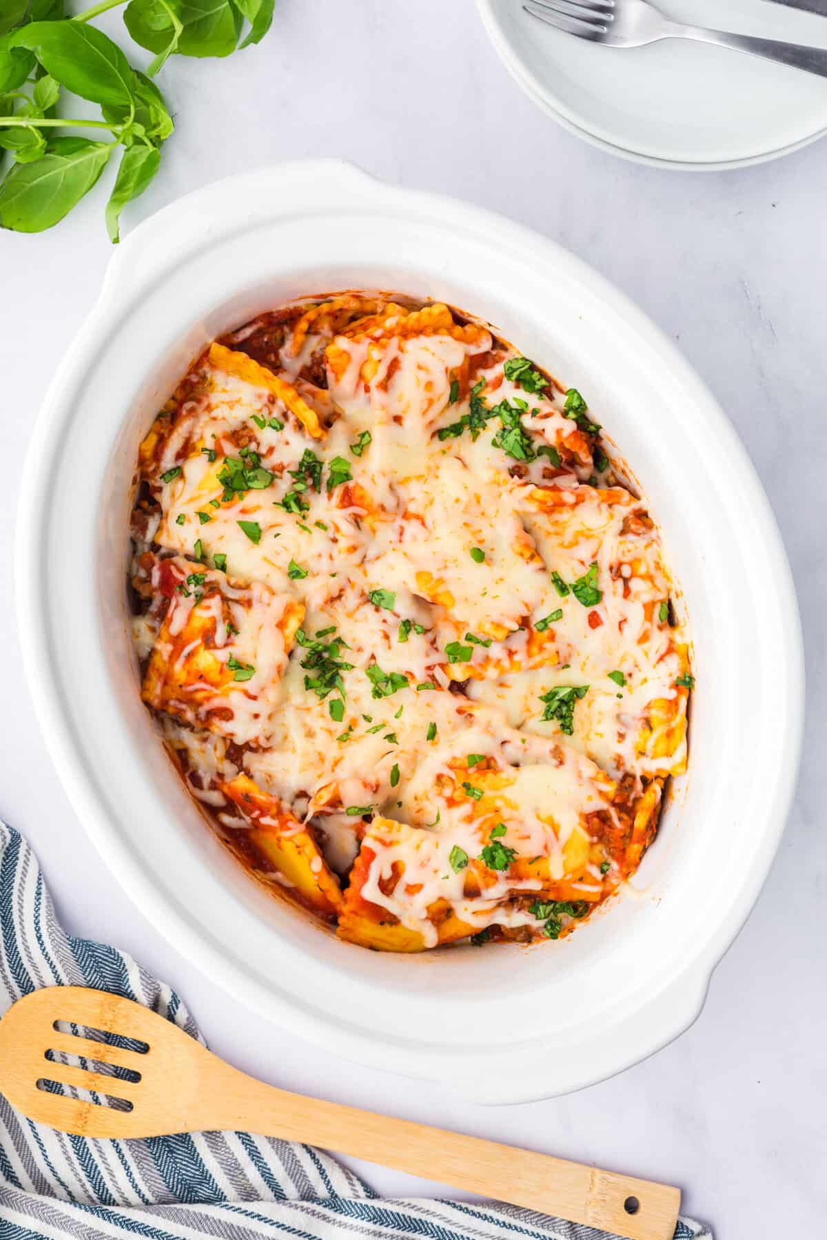 Overhead view of a wooden spoon in a slow cooker full of cheesy ravioli topped with basil.
