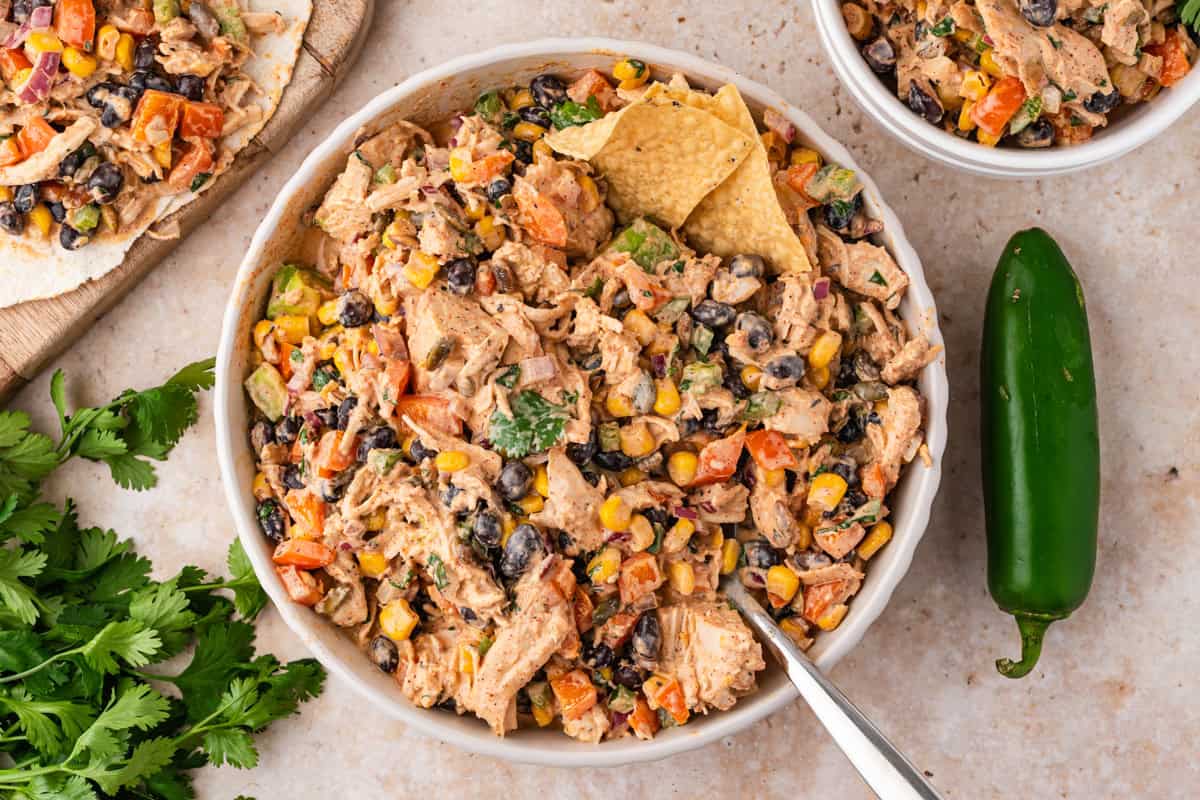Overhead bowl of chicken salad with corn, black beans, peppers, a tortilla chip, and a fresh jalapeno beside it.
