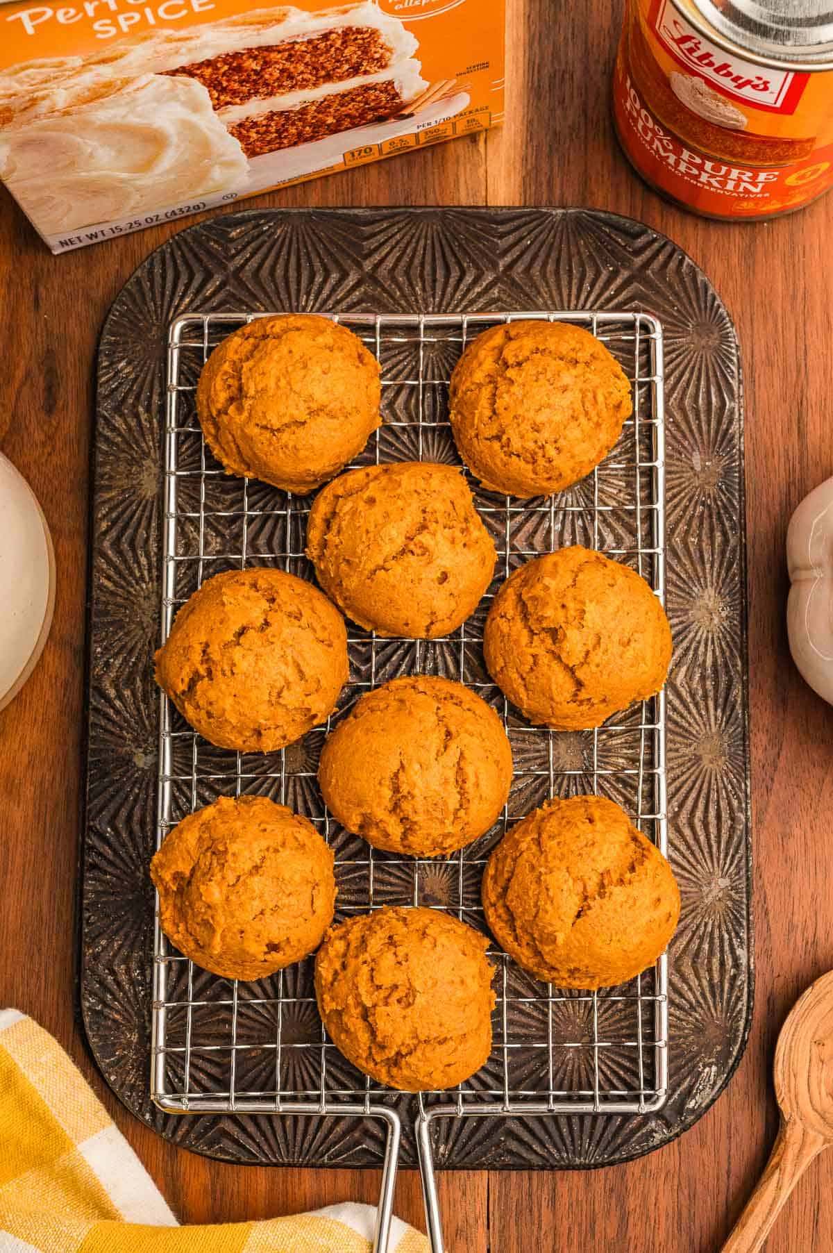 Pumpkin cookies on a wire rack over a dark pan, with a cake mix box, pumpkin can, and wooden spoon nearby.