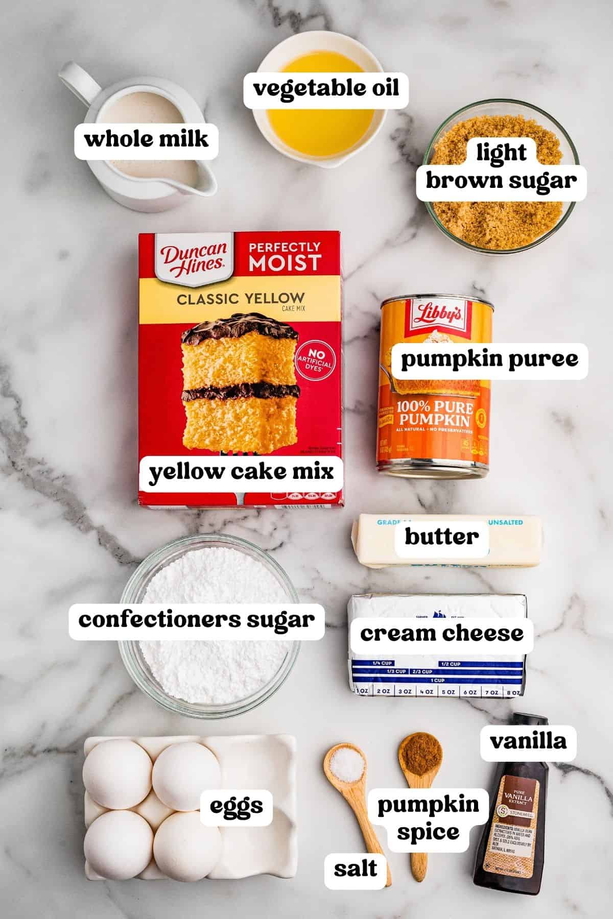 Labeled ingredients on marble: yellow cake mix, pumpkin puree, eggs, brown sugar, oil, milk, butter, cream cheese, and pumpkin spice.