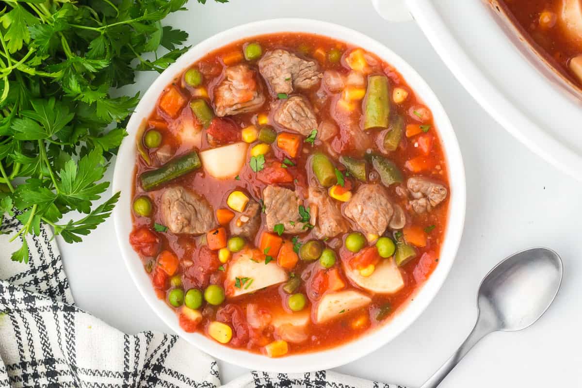 Overhead view of a white bowl of vegetable beef soup with a spoon, parsley and a striped towel beside it.