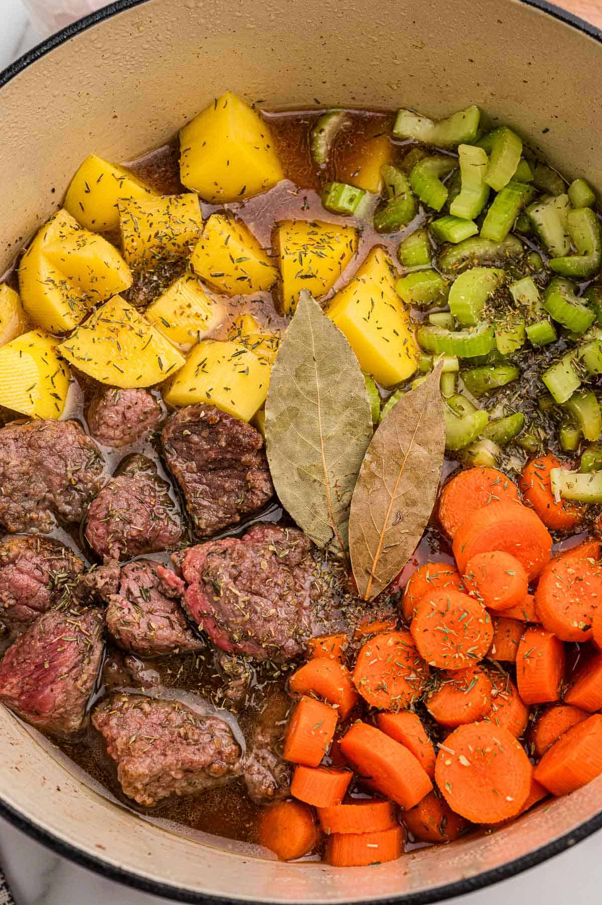 Browned beef, potatoes, celery, carrots, and two bay leaves arranged in a Dutch oven before simmering.