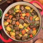 Overhead view of a red Dutch oven filled with beef, potatoes, carrots, celery, and peas.