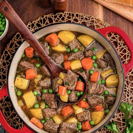 Red Dutch oven of beef stew with a wooden spoon resting inside, bread, butter, and parsley nearby.