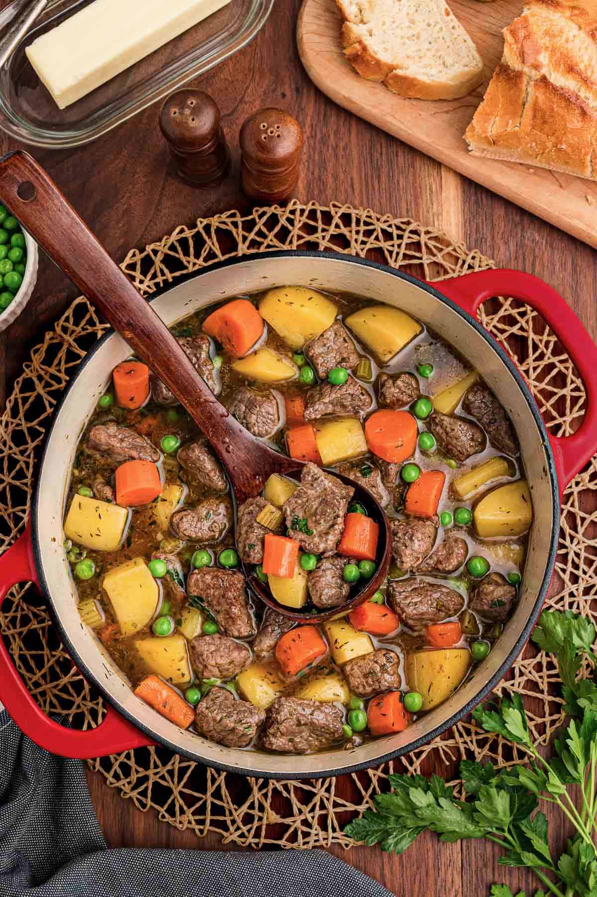 Red Dutch oven of beef stew with a wooden spoon resting inside, bread, butter, and parsley nearby.