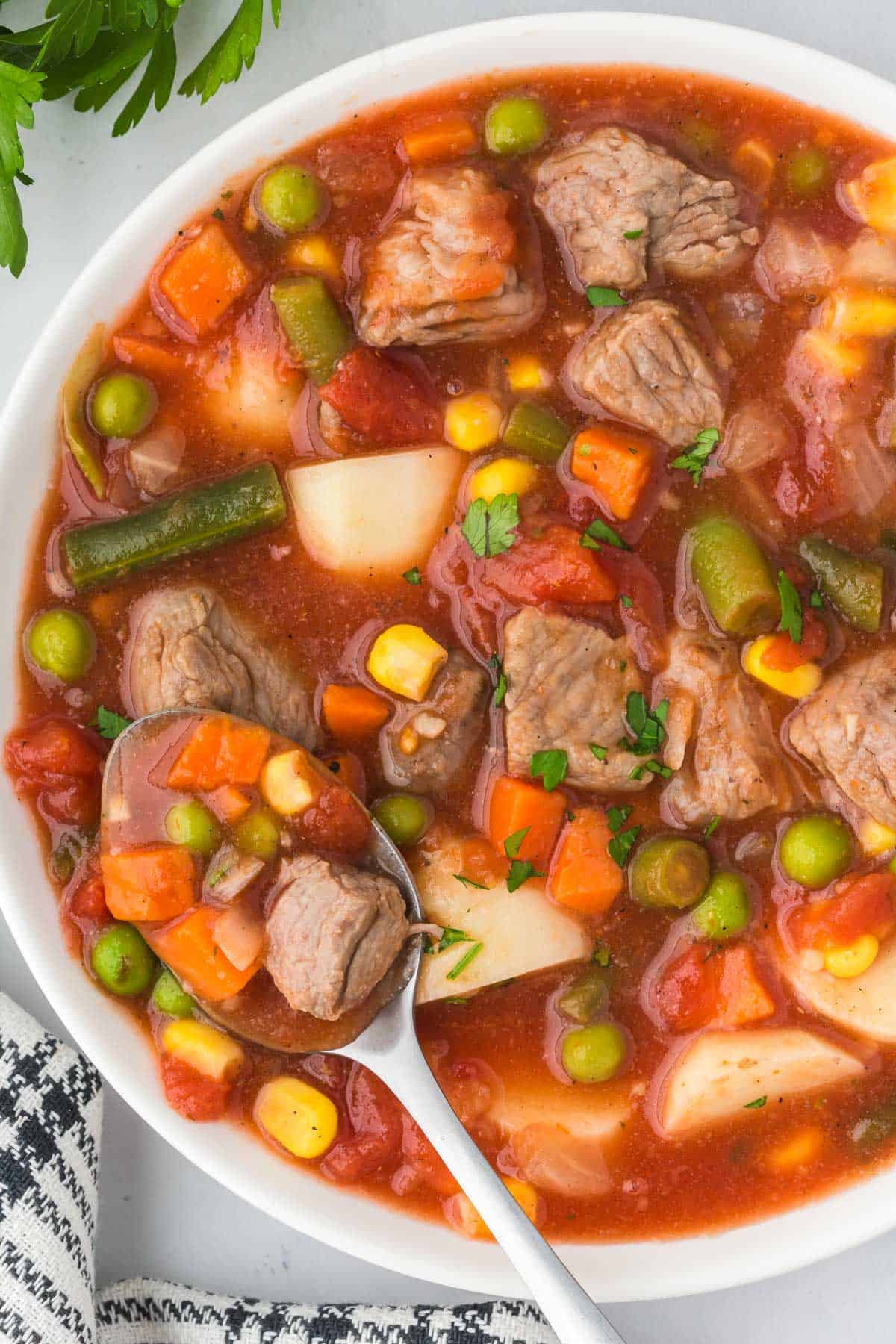 A bowl of vegetable beef soup with a spoon lifting a piece of beef, carrots and peas.