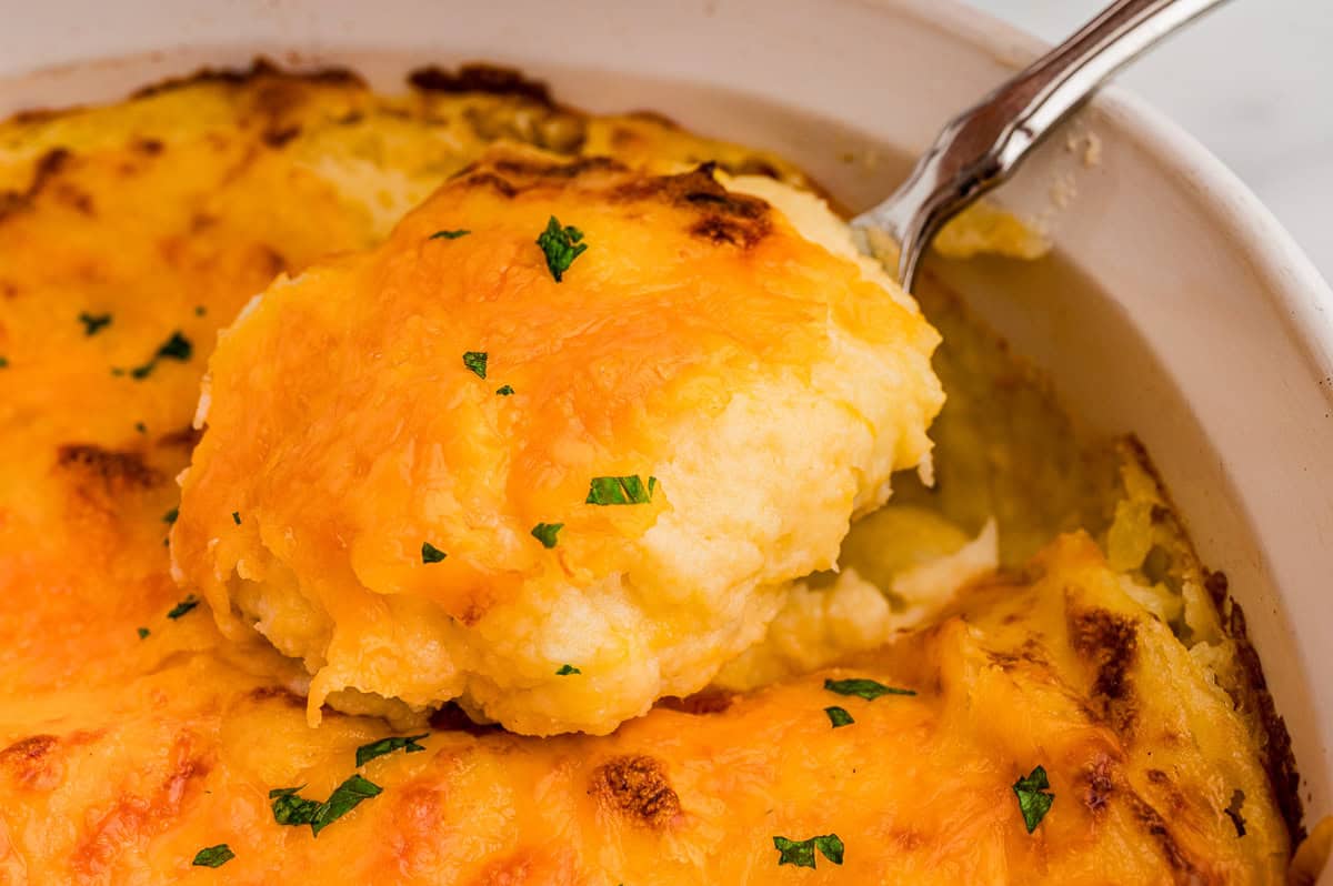 Close-up of a serving spoon scooping creamy potatoes topped with melted, browned cheddar and chopped parsley.