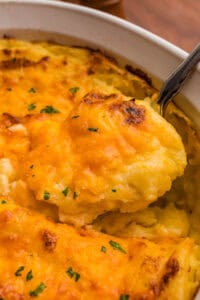 A spoon lifting a scoop of mashed potatoes with a melted, browned cheddar top from a white dish.