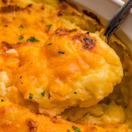 A spoon lifting a scoop of mashed potatoes with a melted, browned cheddar top from a white dish.