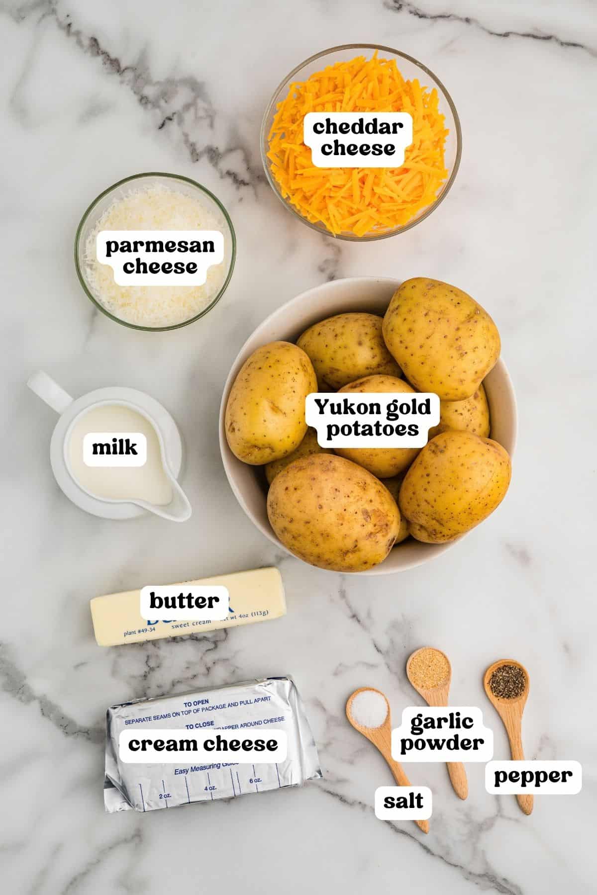 Labeled ingredients on a marble counter: Yukon Gold potatoes, shredded cheddar, Parmesan, milk, butter, cream cheese, garlic powder, salt, and pepper.