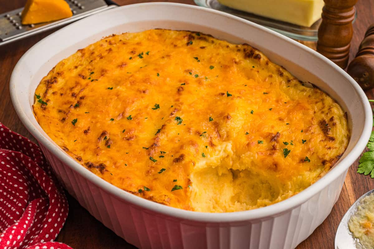 Cheesy mashed potatoes baked in a white oval casserole dish with a golden cheddar top and one scoop taken out.