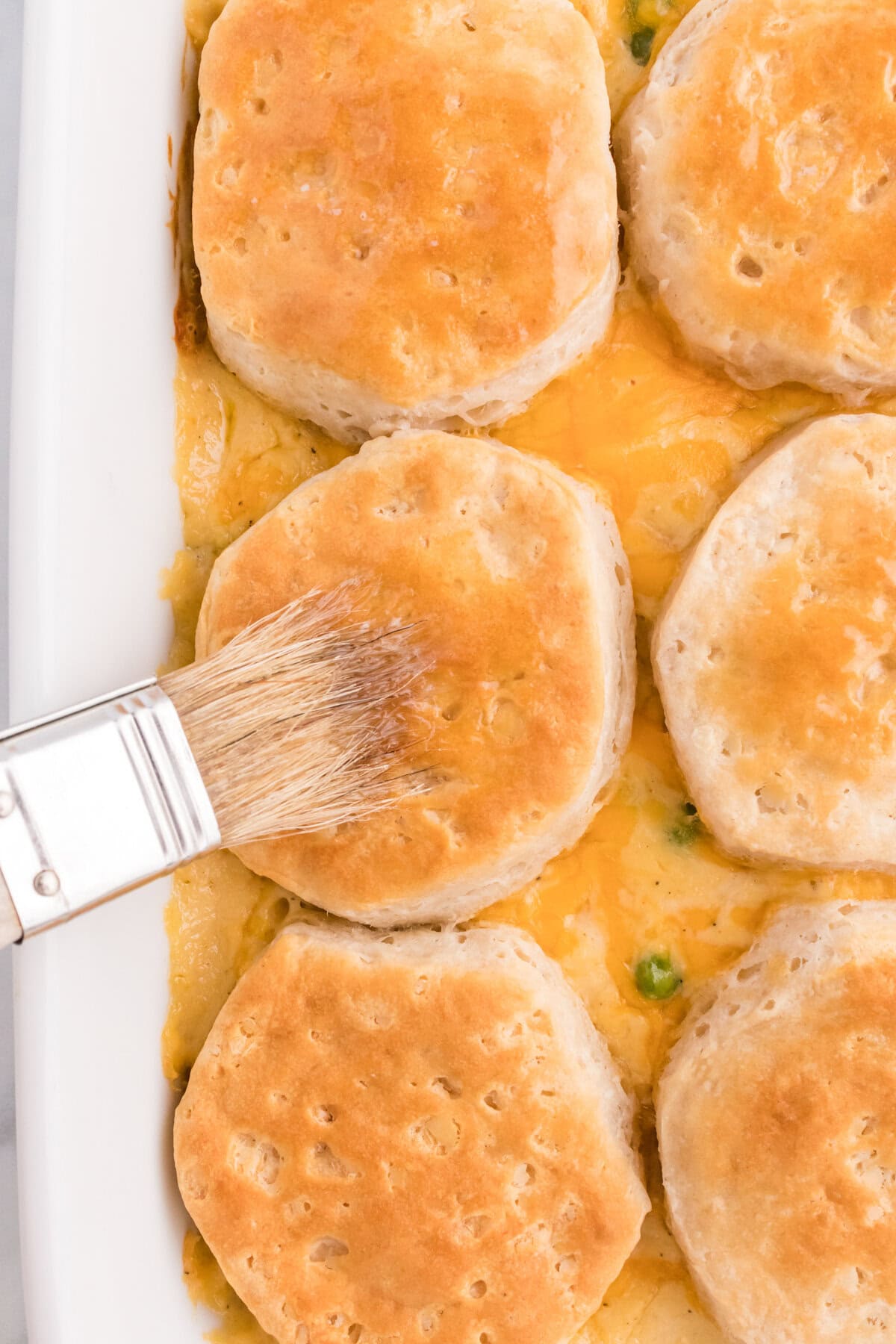 A pastry brush spreading melted butter over golden baked biscuits in a white casserole dish.