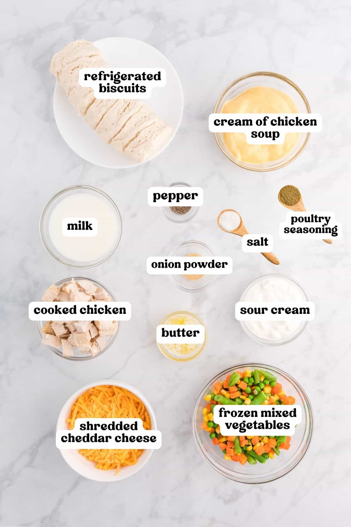 Labeled bowls of cooked chicken, cheddar, frozen mixed vegetables, cream of chicken soup, sour cream, milk, butter, onion powder, poultry seasoning, salt, pepper, and refrigerated biscuits on marble.
