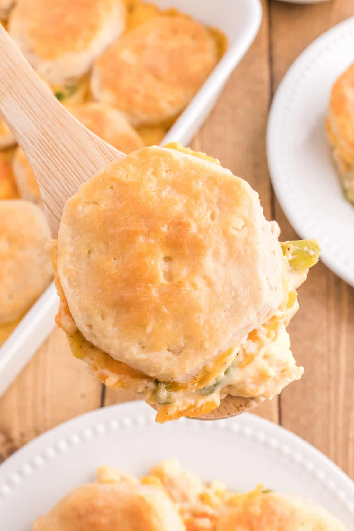A wooden spoon lifting a biscuit topped portion of creamy chicken and vegetables from the casserole dish.