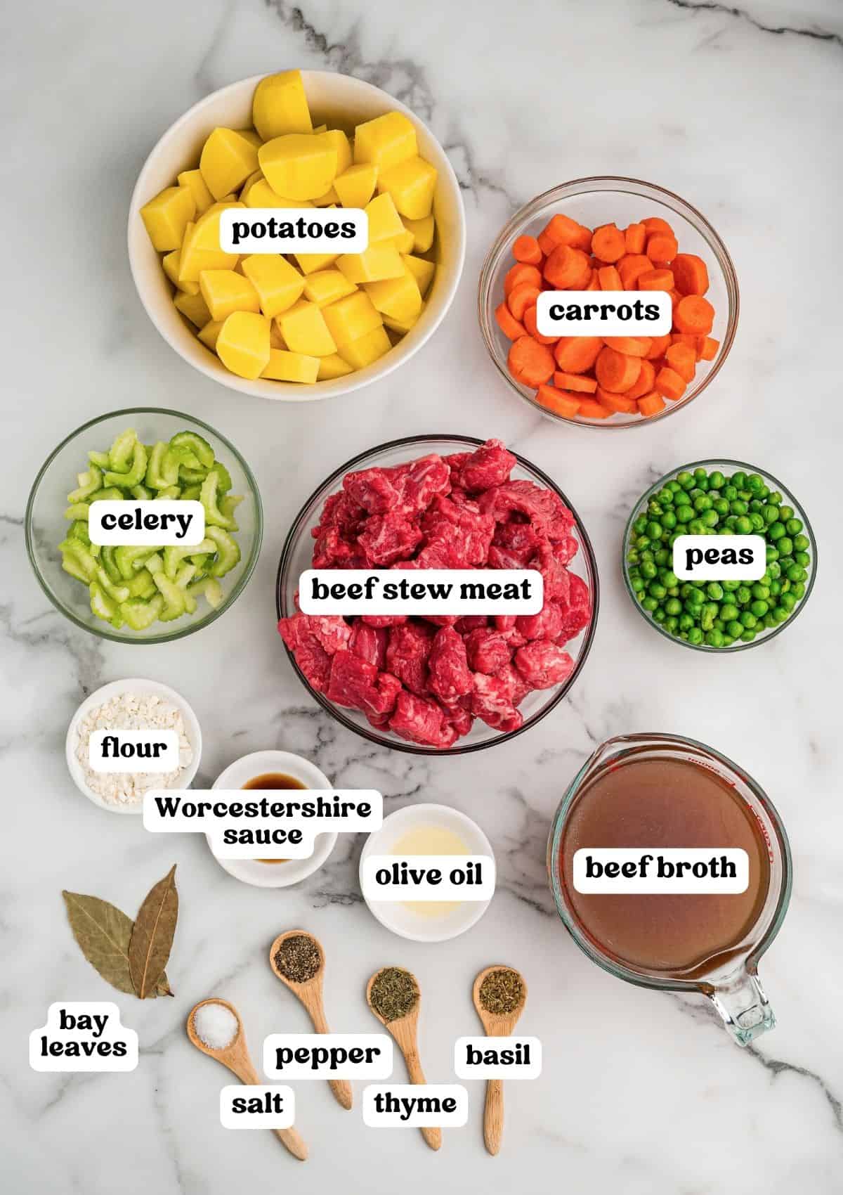 Beef stew meat, potatoes, carrots, celery, and peas in bowls on marble, with flour, olive oil, Worcestershire sauce, broth, and dried spices.