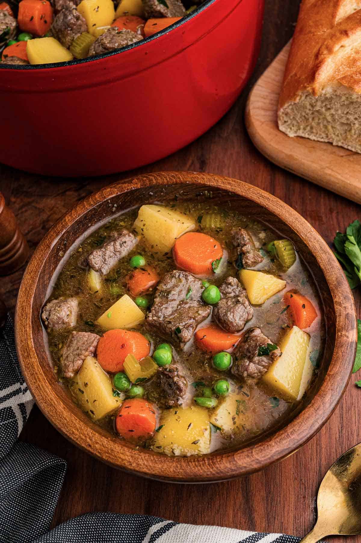 Beef stew served in a wooden bowl with a gold spoon, crusty bread and the pot behind it.