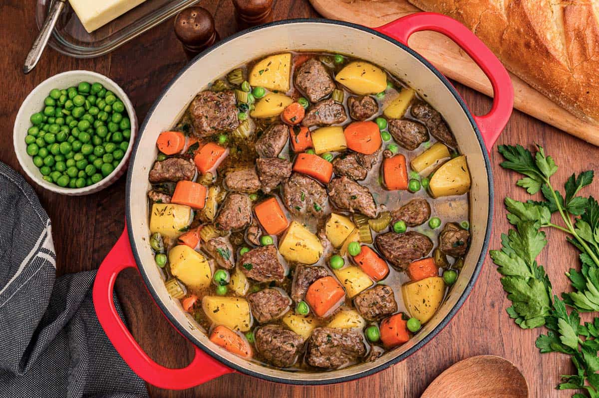 Easy beef stew with potatoes, carrots, celery, and peas in a red Dutch oven beside crusty bread.