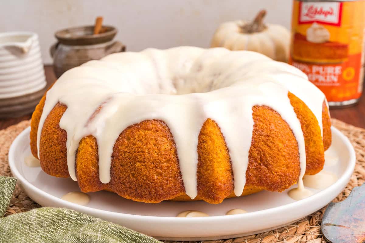 Whole glazed pumpkin bundt cake on a white plate over a woven mat with a green napkin.