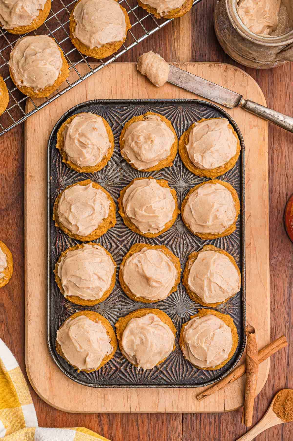 Overhead view of twelve frosted pumpkin cookies in a metal pan on a wooden board with cinnamon sticks.
