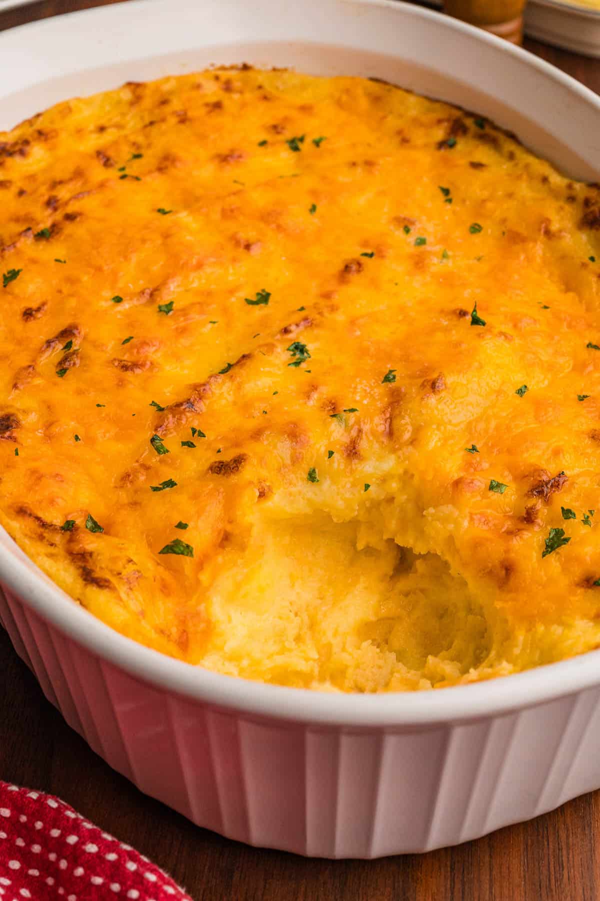 White oval baking dish of mashed potatoes with a golden cheddar crust and a scoop removed, next to a red polka dot towel.