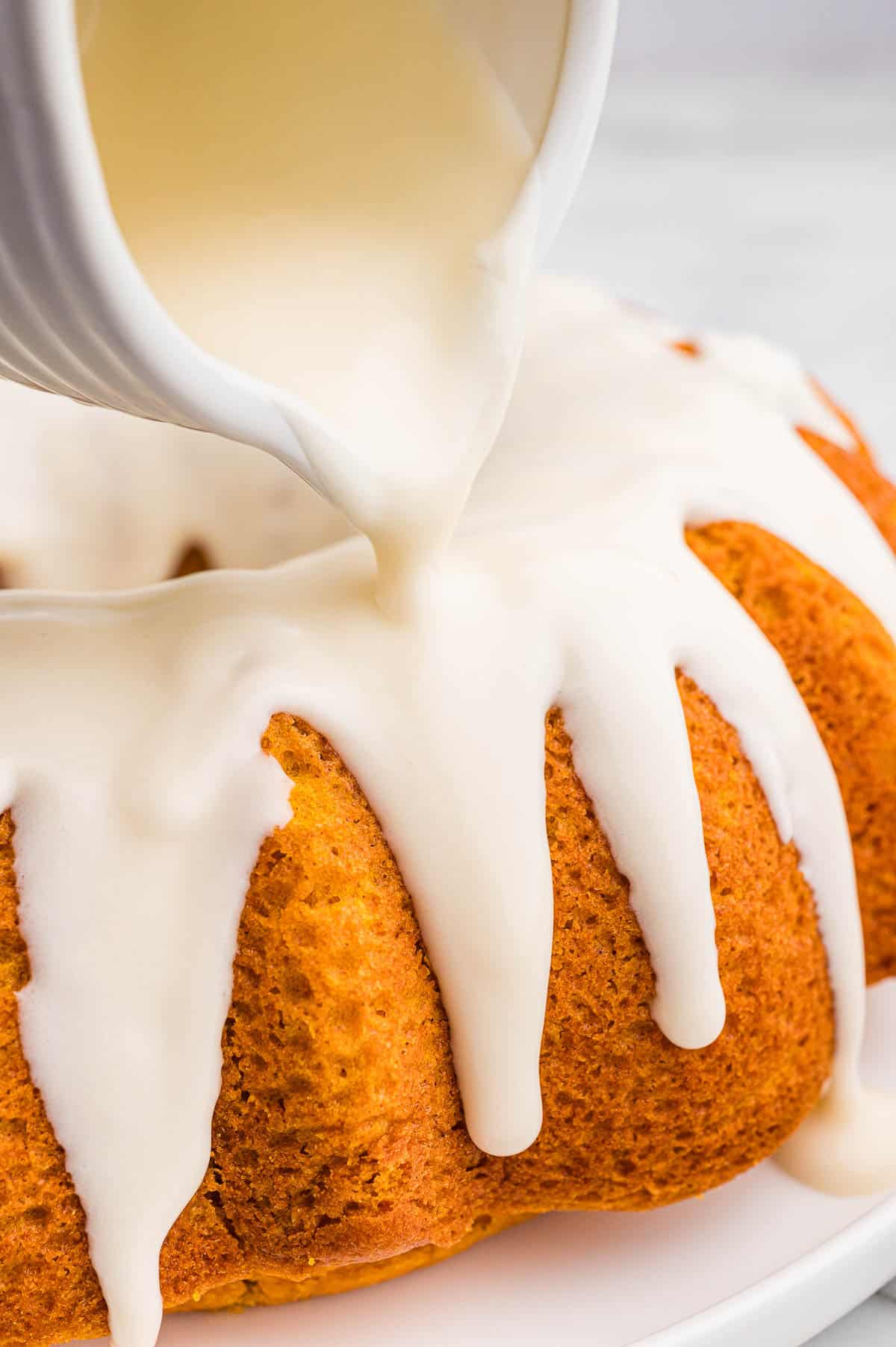 Cream cheese glaze being poured from a white pitcher over a pumpkin bundt cake and dripping down the ridges.