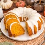 Glazed pumpkin bundt cake cut into slices on a white plate with small pumpkins and a cinnamon crock behind it.
