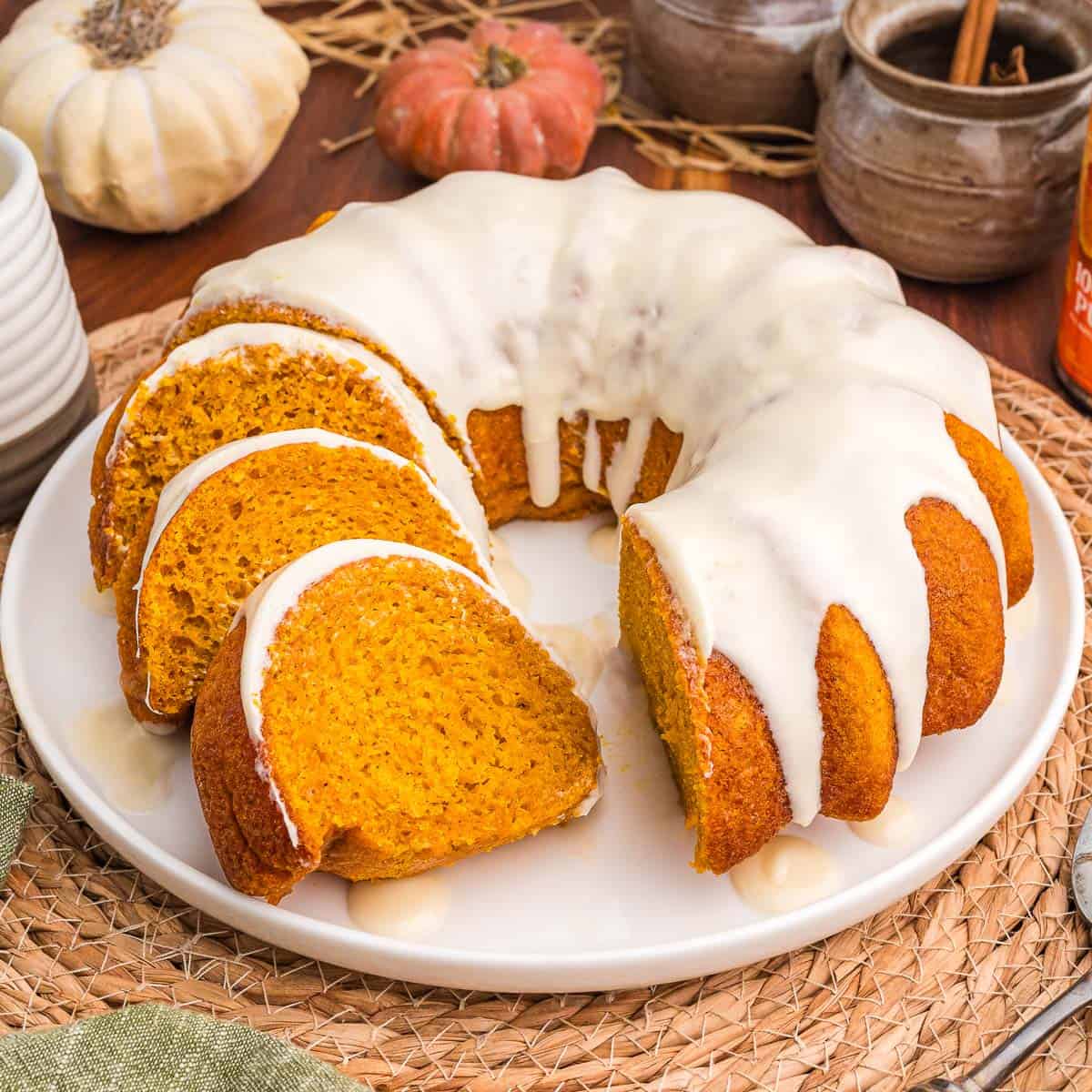 Glazed pumpkin bundt cake cut into slices on a white plate with small pumpkins and a cinnamon crock behind it.