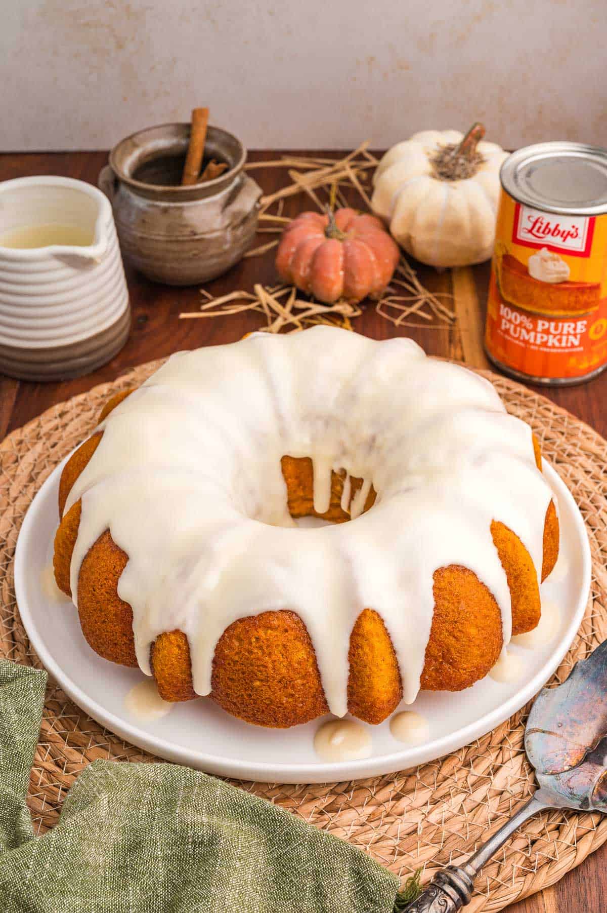 Pumpkin bundt cake with cake mix on a white plate with cream cheese glaze, small pumpkins and a can of pumpkin behind it.