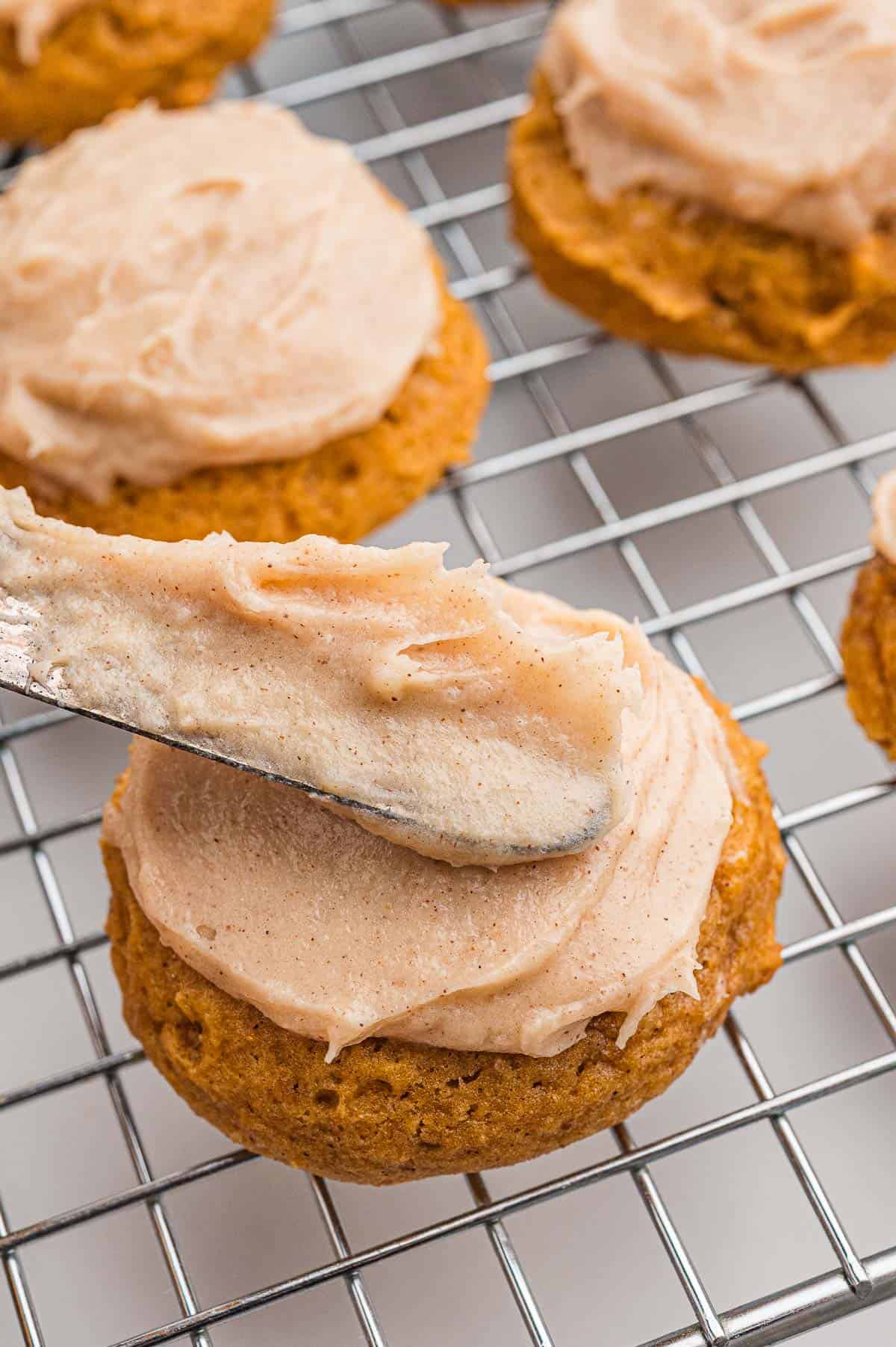 An offset spatula spreading cinnamon cream cheese frosting onto a cookie on a wire cooling rack.