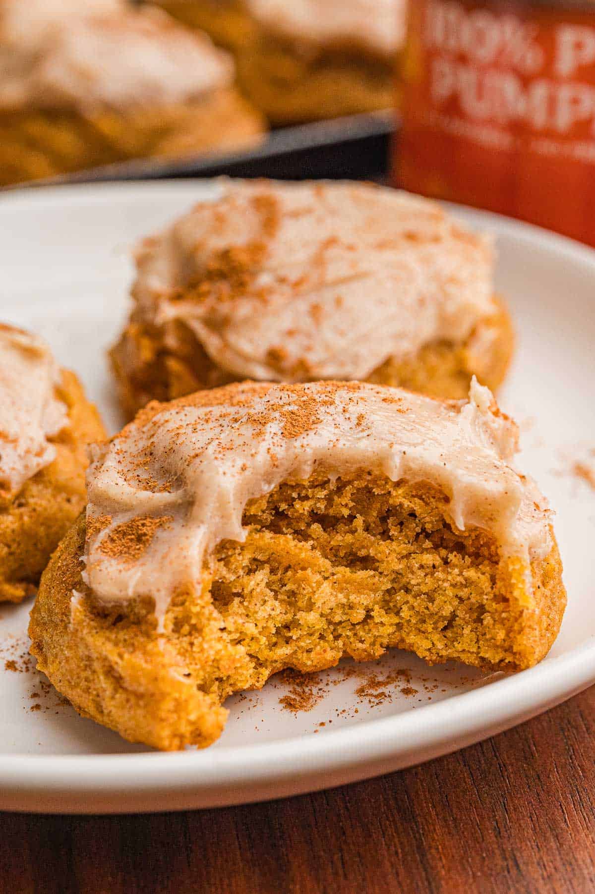 A frosted pumpkin cookie with a bite taken on a white plate, dusted with cinnamon.
