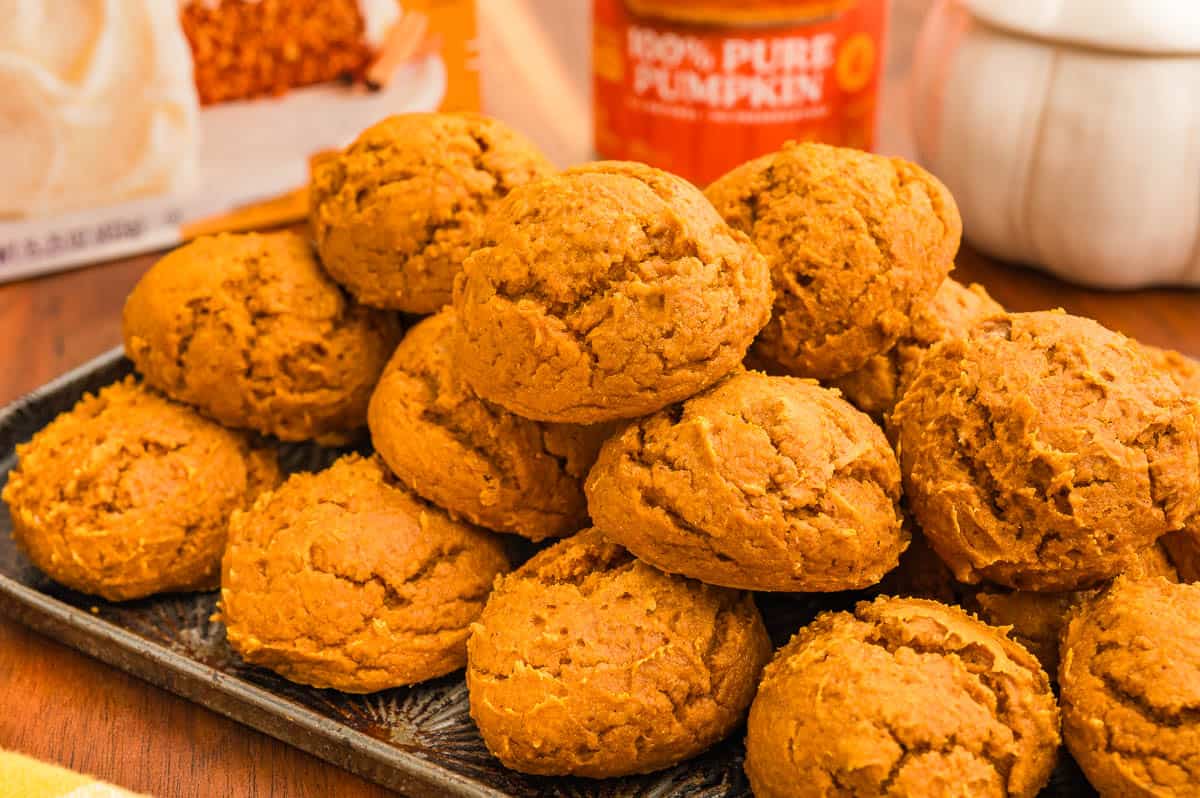 Pumpkin cookies with cake mix stacked on a dark tray with a spice cake box and a can of pure pumpkin behind.