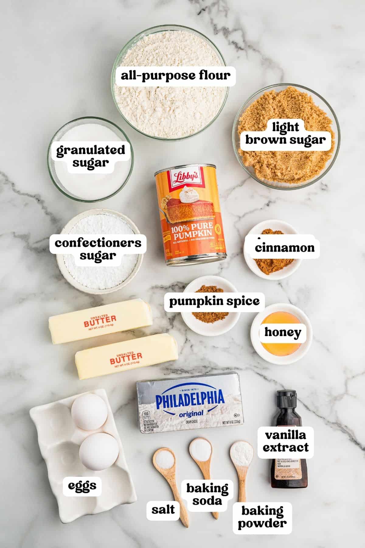 Ingredients on a marble counter: flour, sugars, pumpkin puree, spices, honey, butter, cream cheese, eggs, vanilla, salt, and leaveners.