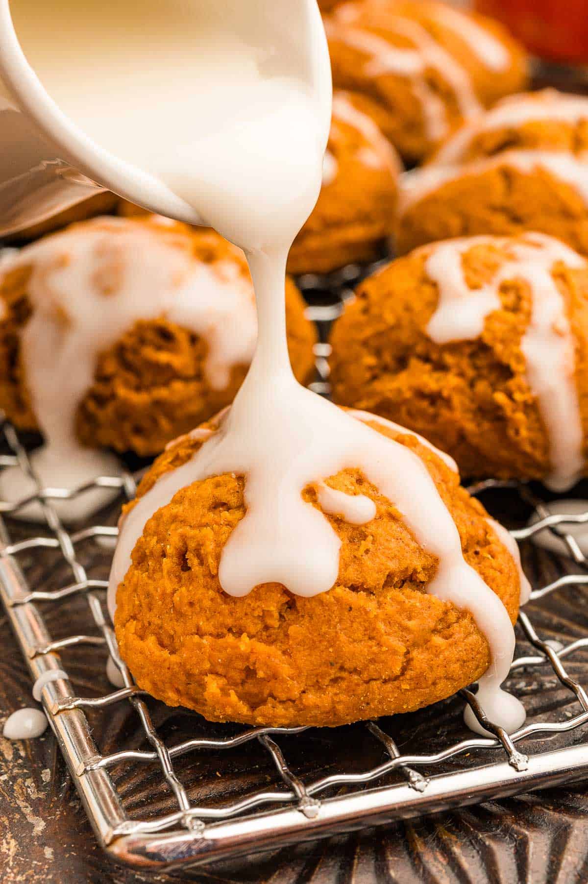 White glaze being poured from a small pitcher over a pumpkin cookie on a wire rack.