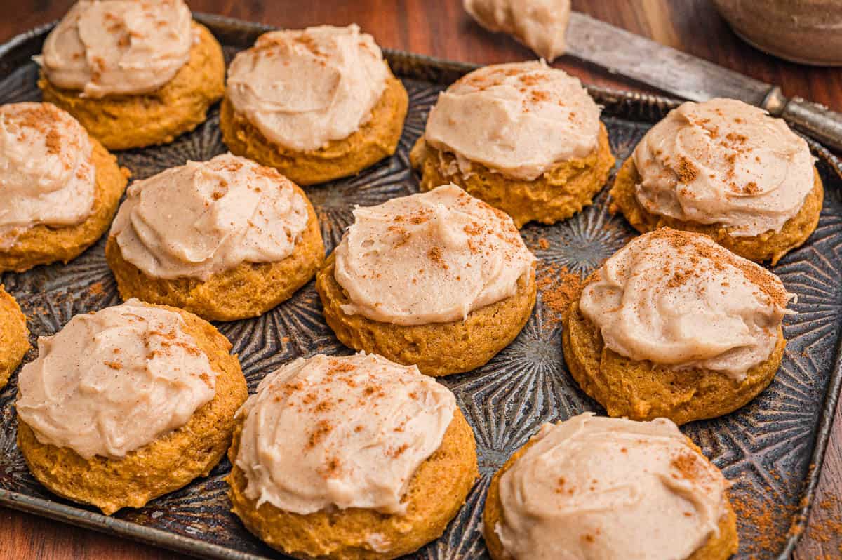 Frosted pumpkin cookies dusted with cinnamon on a dark metal pan with a frosting knife behind.