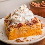 Pumpkin dump cake square on a white plate with whipped cream, cinnamon, and pecans scattered around it.