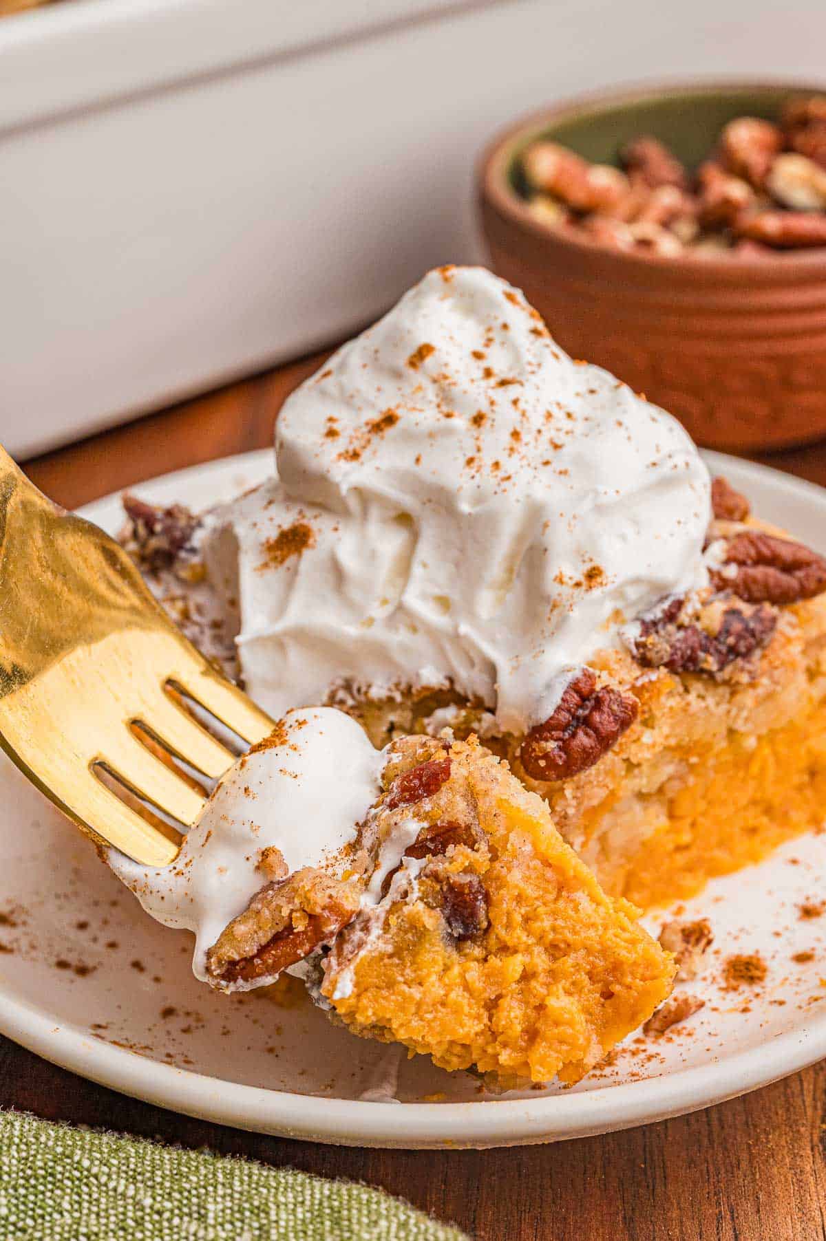 A gold fork lifting a bite of pumpkin cake with whipped cream from a plated square.
