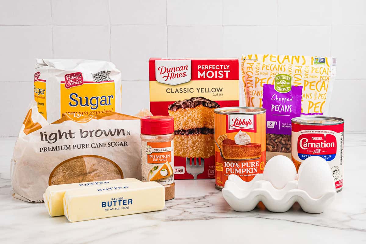 Ingredients on a marble counter: granulated sugar, light brown sugar, pumpkin pie spice, butter, yellow cake mix, chopped pecans, canned pumpkin, evaporated milk, and eggs.