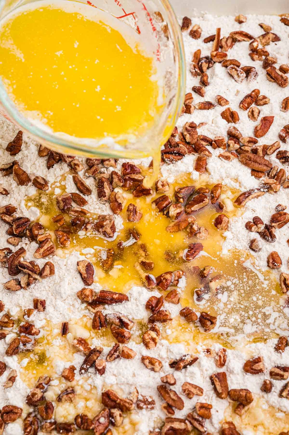 Melted butter being poured from a glass measuring cup over dry cake mix and chopped pecans in a baking dish.
