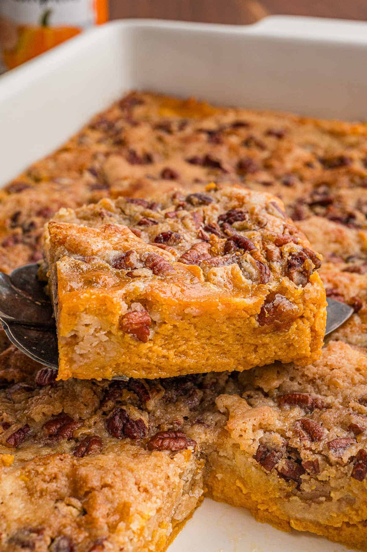A square lifted on a serving spatula showing the orange pumpkin layer under a pecan crumb topping.