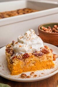 Pumpkin dump cake square on a white plate with whipped cream, cinnamon, and pecans scattered around it and full dump cake behind it.