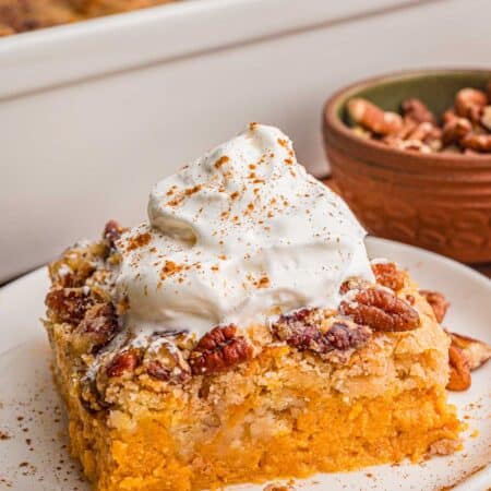 Pumpkin dump cake square on a white plate with whipped cream, cinnamon, and pecans scattered around it and full dump cake behind it.