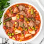 Square overhead view of a white bowl filled with beef, potatoes, green beans, corn and peas in tomato broth.