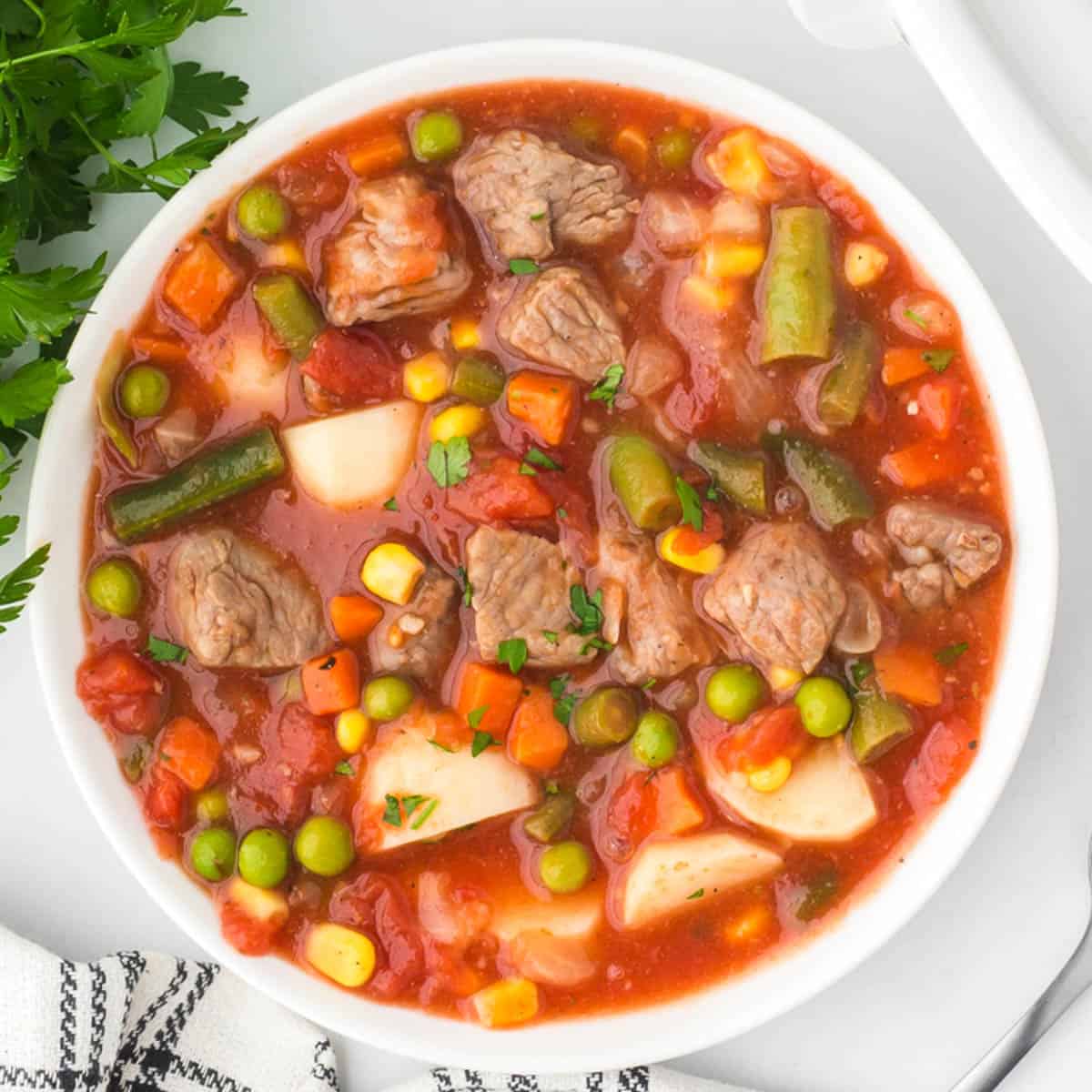 Square overhead view of a white bowl filled with beef, potatoes, green beans, corn and peas in tomato broth.