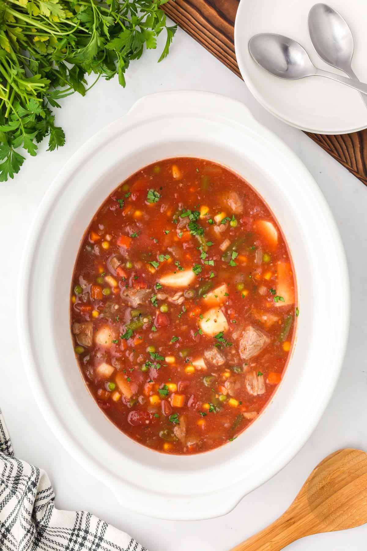 Overhead view of slow cooker vegetable beef soup in a white crockpot with parsley, bowls and spoons alongside.