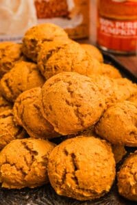 A pile of pumpkin cookies on a dark tray with a spice cake box and pumpkin can behind.