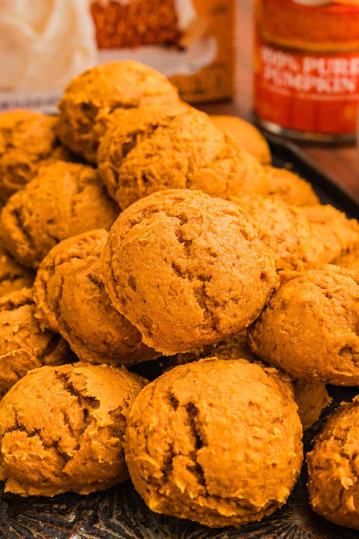 A pile of pumpkin cookies on a dark tray with a spice cake box and pumpkin can behind.