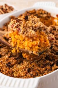A wooden spoon lifting a scoop of mashed sweet potato filling and pecan streusel out of the baking dish.